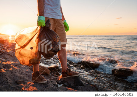A male volunteer holding two polyethilene bag with garbage. Close up. In the background the sea and the sunset. Copy space. The concept of environmental protection and Earth day A male volunteer holding two polyethilene bag with garbage. Close up. In the background the sea and the sunset. Copy space. The concept of environmental protection and Earth day 140146502