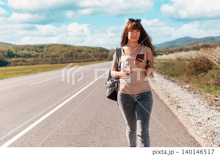 Portrait of pretty young woman with backpack and tattoed hand standing on the empty road and using a smartphone. Copy space. Concept of local traveling and online GPS Portrait of pretty young woman with backpack and tattoed hand standing on the empty road and using a smartphone. Copy space. Concept of local traveling and online GPS 140146517