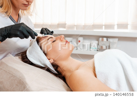 Professional cosmetology. Beautician in rubber gloves cleans the client's forehead using a hydra-peeling device. Side view. Concept of beauty procedures 140146558