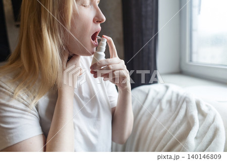 Blonde woman using throat spray to relieve pain. Close up of female treating sore throat or seasonal illness with medication at home. Blonde woman using throat spray to relieve pain. Close up of female treating sore throat or seasonal illness with medication at home. 140146809