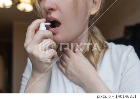 Blonde woman using throat spray to relieve pain. Close up of female treating sore throat or seasonal illness with medication at home. Blonde woman using throat spray to relieve pain. Close up of female treating sore throat or seasonal illness with medication at home. 140146811