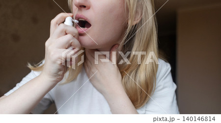Blonde woman using throat spray to relieve pain. Close up of female treating sore throat or seasonal illness with medication at home. Blonde woman using throat spray to relieve pain. Close up of female treating sore throat or seasonal illness with medication at home. 140146814