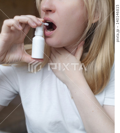Blonde woman using throat spray to relieve pain. Close up of female treating sore throat or seasonal illness with medication at home. Blonde woman using throat spray to relieve pain. Close up of female treating sore throat or seasonal illness with medication at home. 140146815
