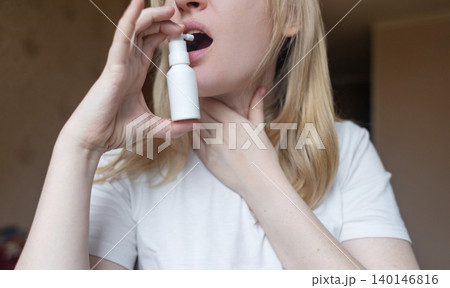 Blonde woman using throat spray to relieve pain. Close up of female treating sore throat or seasonal illness with medication at home. 140146816