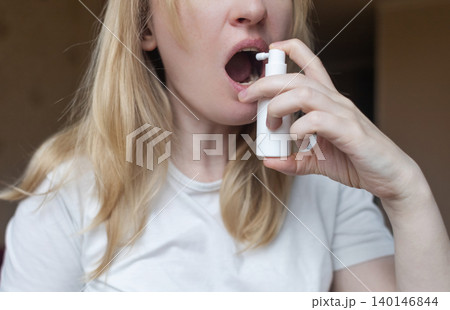 Blonde woman using throat spray to relieve pain. Close up of female treating sore throat or seasonal illness with medication at home. Blonde woman using throat spray to relieve pain. Close up of female treating sore throat or seasonal illness with medication at home. 140146844