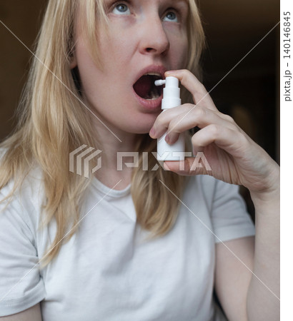 Blonde woman using throat spray to relieve pain. Close up of female treating sore throat or seasonal illness with medication at home. Blonde woman using throat spray to relieve pain. Close up of female treating sore throat or seasonal illness with medication at home. 140146845