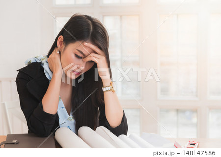 Young asian woman architect holding forehead and neck while working on project at office desk. She is tired and tired. Young asian woman architect holding forehead and neck while working on project at office desk. She is tired and tired. 140147586