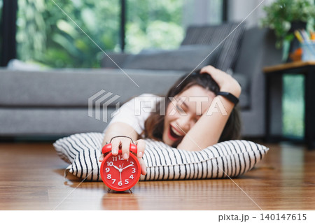 Young asian woman open mouth yawning and reaching over to turning off alarm clock while lying on the floor in living room at home. Young asian woman open mouth yawning and reaching over to turning off alarm clock while lying on the floor in living room at home. 140147615