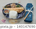 A grilled Shima Hokke fillet sits on a deep blue patterned plate, paired with a mound of white rice and a small dipping bowl. Three traditional sides hijiki seaweed, simmered roots, and greens line 140147690