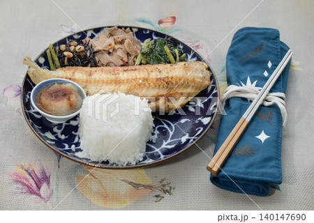 A grilled Shima Hokke fillet sits on a deep blue patterned plate, paired with a mound of white rice and a small dipping bowl. Three traditional sides hijiki seaweed, simmered roots, and greens line A grilled Shima Hokke fillet sits on a deep blue patterned plate, paired with a mound of white rice and a small dipping bowl. Three traditional sides hijiki seaweed, simmered roots, and greens line 140147690