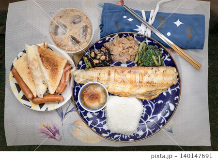 A blue patterned plate holds a grilled fish fillet, white rice, grated radish, and three sides: hijiki seaweed, simmered roots, and greens. To the left, a smaller plate has toast triangles and three 140147691