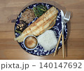 A grilled golden-brown fish fillet rests diagonally on a patterned deep-blue ceramic plate over a wooden table. It is accompanied by a square-molded portion of white rice, a small condiment bowl and 140147692