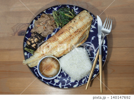 A grilled golden-brown fish fillet rests diagonally on a patterned deep-blue ceramic plate over a wooden table. It is accompanied by a square-molded portion of white rice, a small condiment bowl and A grilled golden-brown fish fillet rests diagonally on a patterned deep-blue ceramic plate over a wooden table. It is accompanied by a square-molded portion of white rice, a small condiment bowl and 140147692