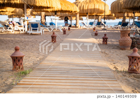Wooden boardwalk leads across a sandy beach toward straw umbrellas sunbeds and the sea in a resort recreation area. Beach leisure vacation tourism resort lifestyle 140149309