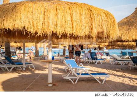 Straw beach umbrellas provide shade above blue sunbeds on a sandy beach with vacationers relaxing near the shoreline. Beach leisure vacation tourism resort lifestyle 140149310