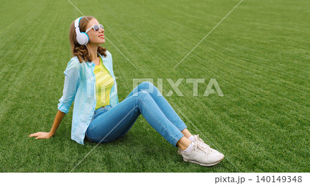 Happy smiling young woman listening to music in headphones while lying on grass in summer park 140149348