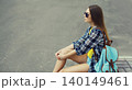 Stylish teenager girl with backpack, young woman sitting on city street on a summer day 140149461