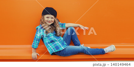 Happy cheerful kid girl dancer with curly hair, joyful child dancing in the city on orange wall Happy cheerful kid girl dancer with curly hair, joyful child dancing in the city on orange wall 140149464
