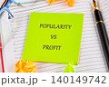A bright green sticky note reads Popularity vs Profit, sitting on lined paper amid colorful crumpled papers and sleek writing tools, inspiring business ideas 140149742