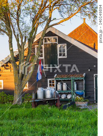 Traditional wooden Dutch farmhouse with national flag, green grass and warm sunset light, representing rural lifestyle, heritage architecture and countryside scenery in the Netherlands. 140149894