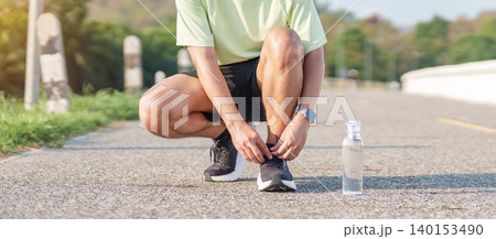 Young Asian man tying running shoes in the park with a reusable water bottle aside. male runner getting ready for jogging and fitness exercise. Healthy lifestyle and sustainability concept Young Asian man tying running shoes in the park with a reusable water bottle aside. male runner getting ready for jogging and fitness exercise. Healthy lifestyle and sustainability concept 140153490