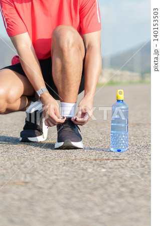 Young Asian man tying running shoes in the park with a reusable water bottle aside. male runner getting ready for jogging and fitness exercise. Healthy lifestyle and sustainability concept 140153503