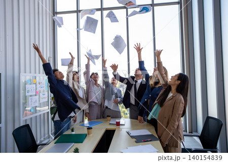 Happy business team celebrating success in office by throwing documents in the air. Corporate achievement, teamwork, motivation, workplace happiness, and startup success concept. 140154839