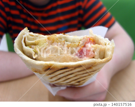 Person in a striped shirt holds a grilled, partially eaten chicken wrap. The close-up reveals fillings of shredded meat, creamy garlic sauce, and fresh tomatoes. A delicious fast food meal Person in a striped shirt holds a grilled, partially eaten chicken wrap. The close-up reveals fillings of shredded meat, creamy garlic sauce, and fresh tomatoes. A delicious fast food meal 140157516