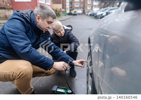 Father and son pumping a car tire with a foot pump on a city street. Caucasian man teaches his boy how to inflate automobile wheels, lifestyle family outdoor activity. Father and son pumping a car tire with a foot pump on a city street. Caucasian man teaches his boy how to inflate automobile wheels, lifestyle family outdoor activity. 140160546