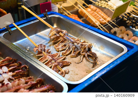 Grilled squid skewers on metal tray for sale at Temple Street Night Market street food stall with diverse seafood and sausages background in Hong Kong Grilled squid skewers on metal tray for sale at Temple Street Night Market street food stall with diverse seafood and sausages background in Hong Kong 140160909