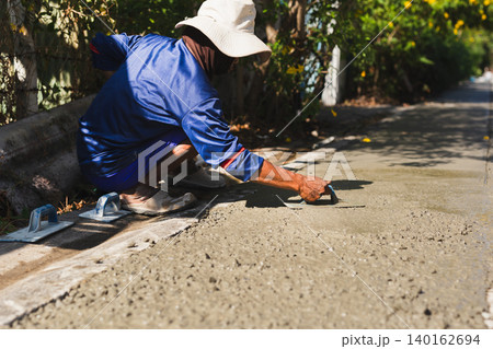 Construction worker crouching on a roadside to smooth wet cement using a hand trowel. Construction worker crouching on a roadside to smooth wet cement using a hand trowel. 140162694
