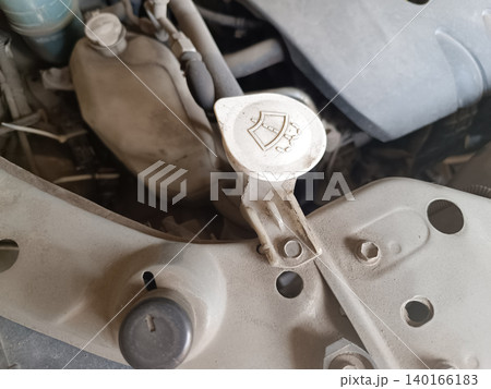 Close-up shot of a dusty car engine bay, focusing on the white plastic cap of the windshield washer fluid reservoir. The cap features the standard windshield wiper symbol, surrounded by dirty engine p Close-up shot of a dusty car engine bay, focusing on the white plastic cap of the windshield washer fluid reservoir. The cap features the standard windshield wiper symbol, surrounded by dirty engine p 140166183