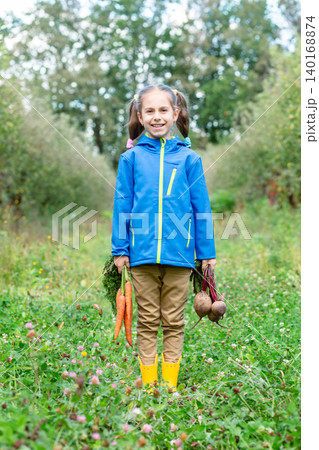 Smiling girl standing orchard holding carrots beets, wearing jacket during autumn harvest outdoors 140168874