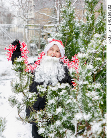 Child dressed as Santa with white beard and red tinsel garland standing outdoors Child dressed as Santa with white beard and red tinsel garland standing outdoors 140169882