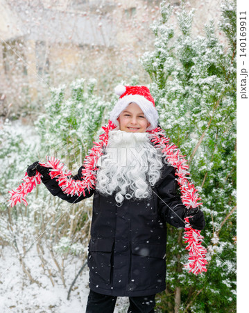 Child dressed as Santa with white beard red tinsel garland standing outdoors in snowy winter garden Child dressed as Santa with white beard red tinsel garland standing outdoors in snowy winter garden 140169911