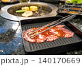 Slices of raw marinated beef in a black tray with metal tongs. Korean BBQ or Japanese yakiniku restaurant scene with a charcoal grill in the background. Fresh meat prepared for grilling. 140170669