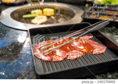 Slices of raw marinated beef in a black tray with metal tongs. Korean BBQ or Japanese yakiniku restaurant scene with a charcoal grill in the background. Fresh meat prepared for grilling. 140170669