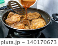Chef pouring savory glaze over pork steaks in a frying pan on a gas stove. Delicious meat marinated and cooked for a gourmet dinner at home. High angle view of professional kitchen cooking process. 140170673