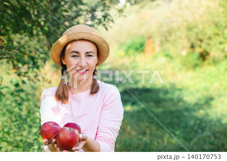 Portrait of woman in hat offering fresh red apples in orchard during warm sunny autumn day outdoors Portrait of woman in hat offering fresh red apples in orchard during warm sunny autumn day outdoors 140170753