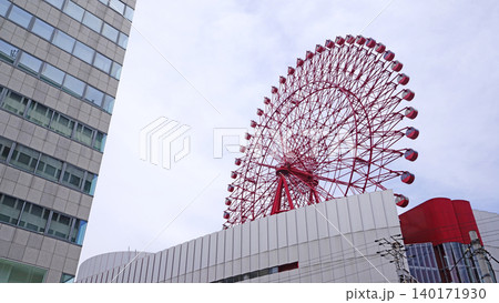 Modern Ferris wheel structure with a tall glass building against a cloudy sky 140171930
