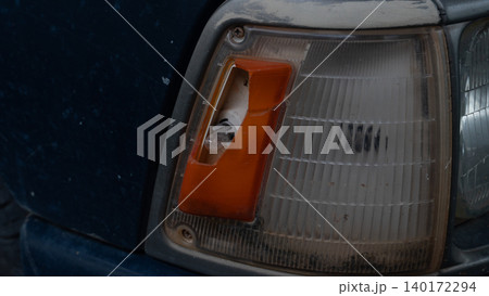 Close up view of a classic car showing a broken orange turn signal indicator lens next to the main headlight housing. The damaged plastic assembly highlights an auto repair need. 140172294