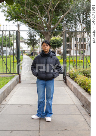 A teenager in a black jacket and blue jeans stands in front of a gate 140175901
