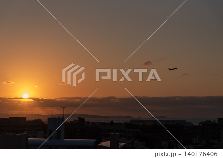 離陸する飛行機と手前に街のシルエットがあるオレンジ色の夕焼け空の太陽と遠くに離島の見える海 140176406