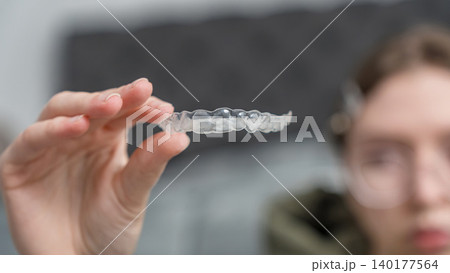 Young woman holds clear dental aligner in hand, showcasing its transparent design against a blurred background of a dental office Young woman holds clear dental aligner in hand, showcasing its transparent design against a blurred background of a dental office 140177564