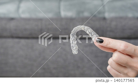 Transparent dental aligner held by a teenager with black nail polish, sitting on a gray couch in a modern living room setting Transparent dental aligner held by a teenager with black nail polish, sitting on a gray couch in a modern living room setting 140177565