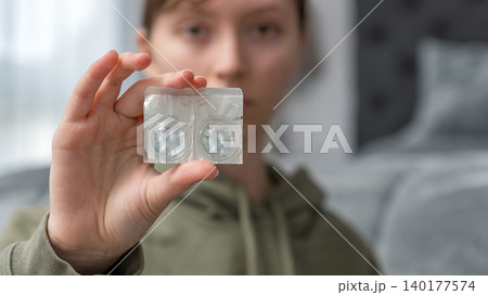 Teenager holding a pack of one-day contact lenses in a bedroom setting, soft lighting and blurred background elements visible 140177574