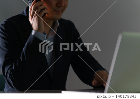 逆光により匿名性が強調されたスーツ姿の男性がパソコンとスマホを操作するサイバー犯罪のイメージ 逆光により匿名性が強調されたスーツ姿の男性がパソコンとスマホを操作するサイバー犯罪のイメージ 140178843