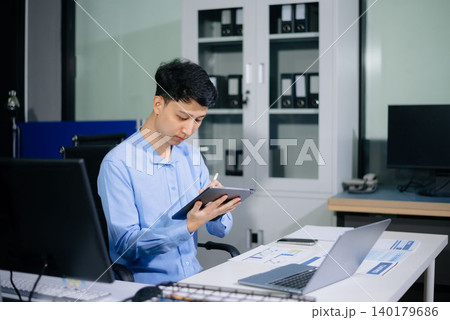 Asian businessman reviewing reports and working on laptop in a bright daytime office, highlighting productivity, business analysis, corporate workflow, and success. Asian businessman reviewing reports and working on laptop in a bright daytime office, highlighting productivity, business analysis, corporate workflow, and success. 140179686