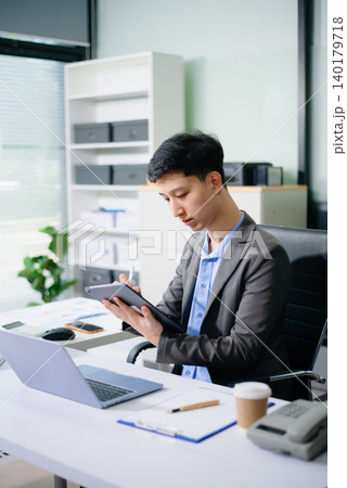 Asian businessman reviewing reports and working on laptop in a bright daytime office, highlighting productivity, business analysis, corporate workflow, and success. Asian businessman reviewing reports and working on laptop in a bright daytime office, highlighting productivity, business analysis, corporate workflow, and success. 140179718