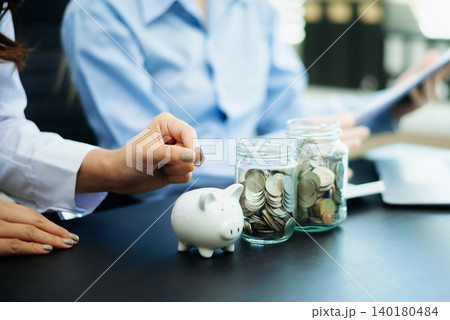 Women saving and counting money with coins, jars, piggy bank, and calculator in office. Finance, budgeting, planning, and smart money Women saving and counting money with coins, jars, piggy bank, and calculator in office. Finance, budgeting, planning, and smart money 140180484
